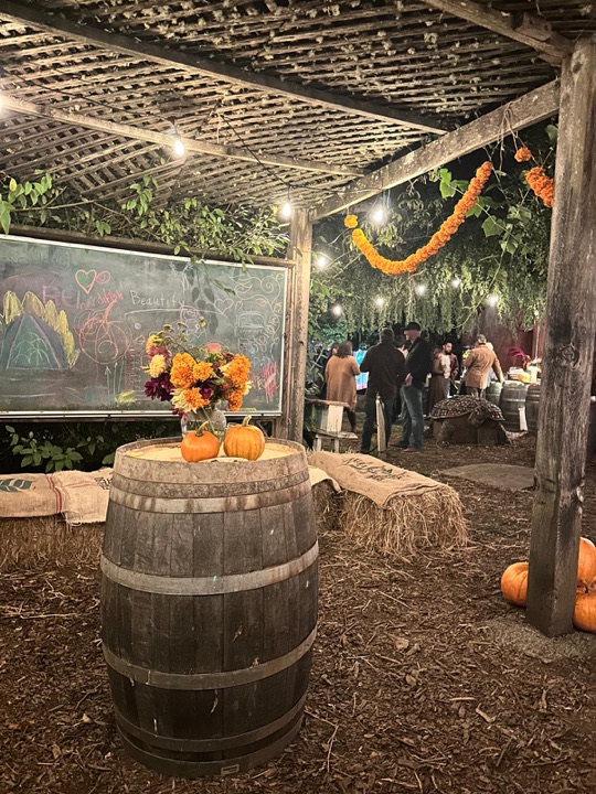 A wine barrel with a jar of marigolds and two small pumpkins; the garden chalkboard and a marigold garland behind.