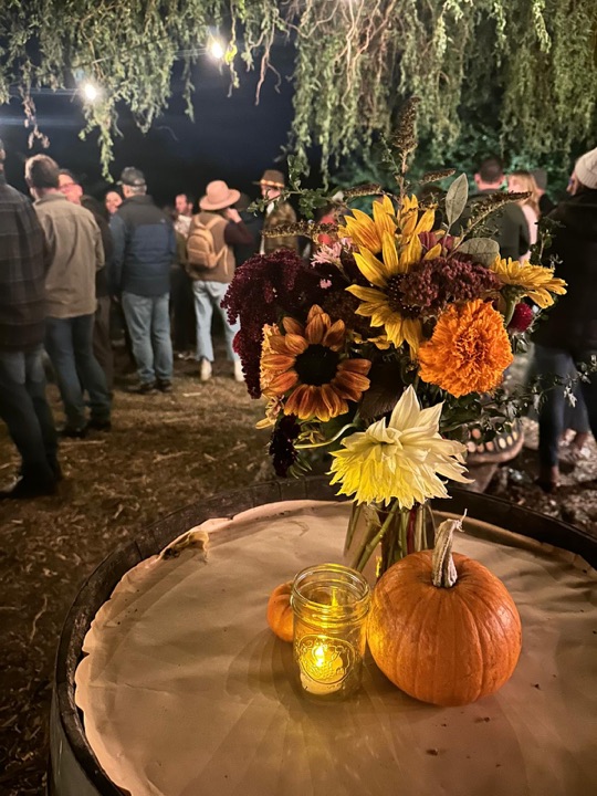 Sunflowers, marigolds and a dahlia in a jar beside a candle and a pumpkin, guests talking behind.