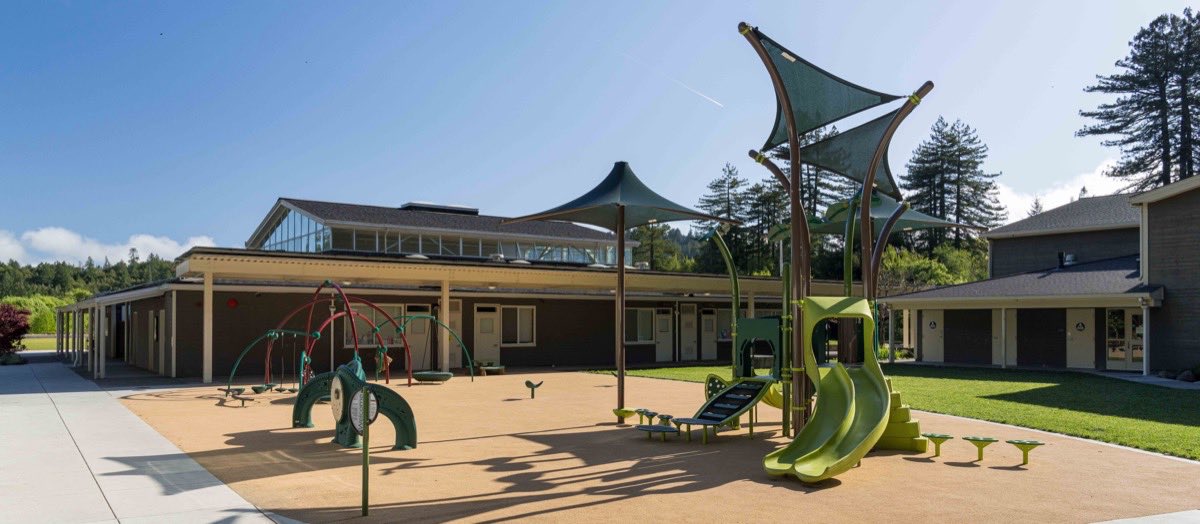 The school playground on a clear day: a green climbing structure with slides under sail shades, swings alongside, and the classroom buildings and redwoods behind.