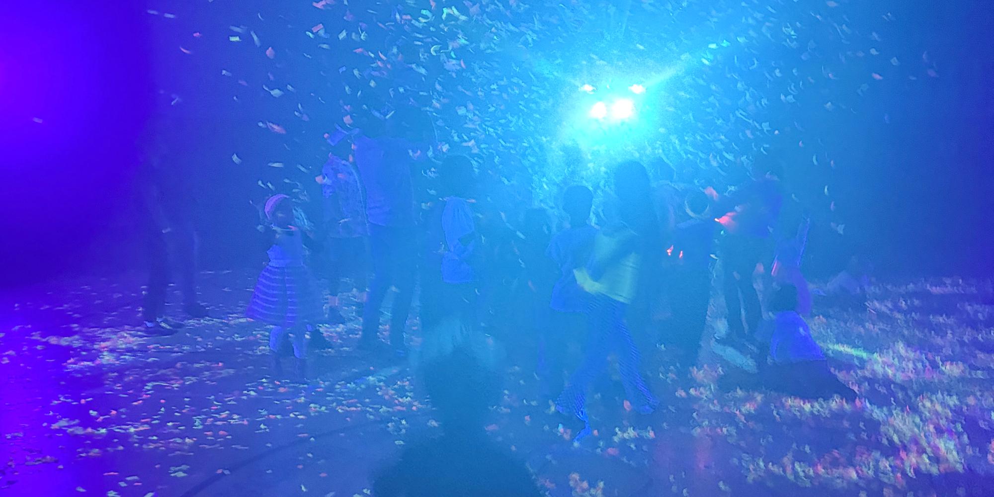 The school gym during last year's Glow Party: confetti in the air, a bright blue light behind the dancers, children in silhouette.