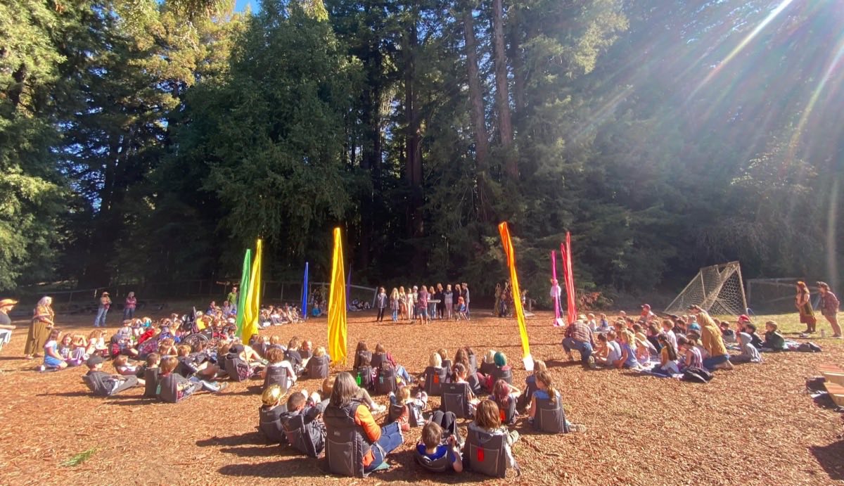 The whole school sitting in a circle on the field, coloured silk banners standing in the middle, tall redwoods and low autumn sun behind.